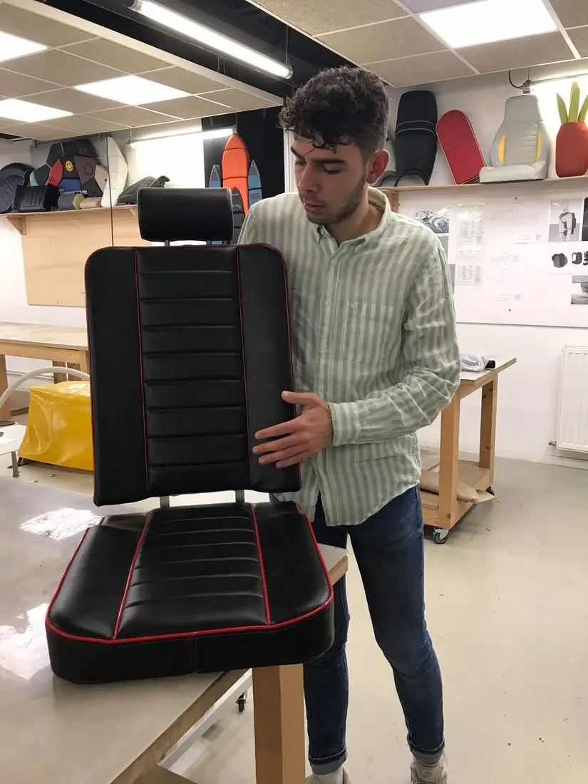 Charles Bailly portant un siège matelassé noir et rouge qu'il vient de terminer, en atelier de formation