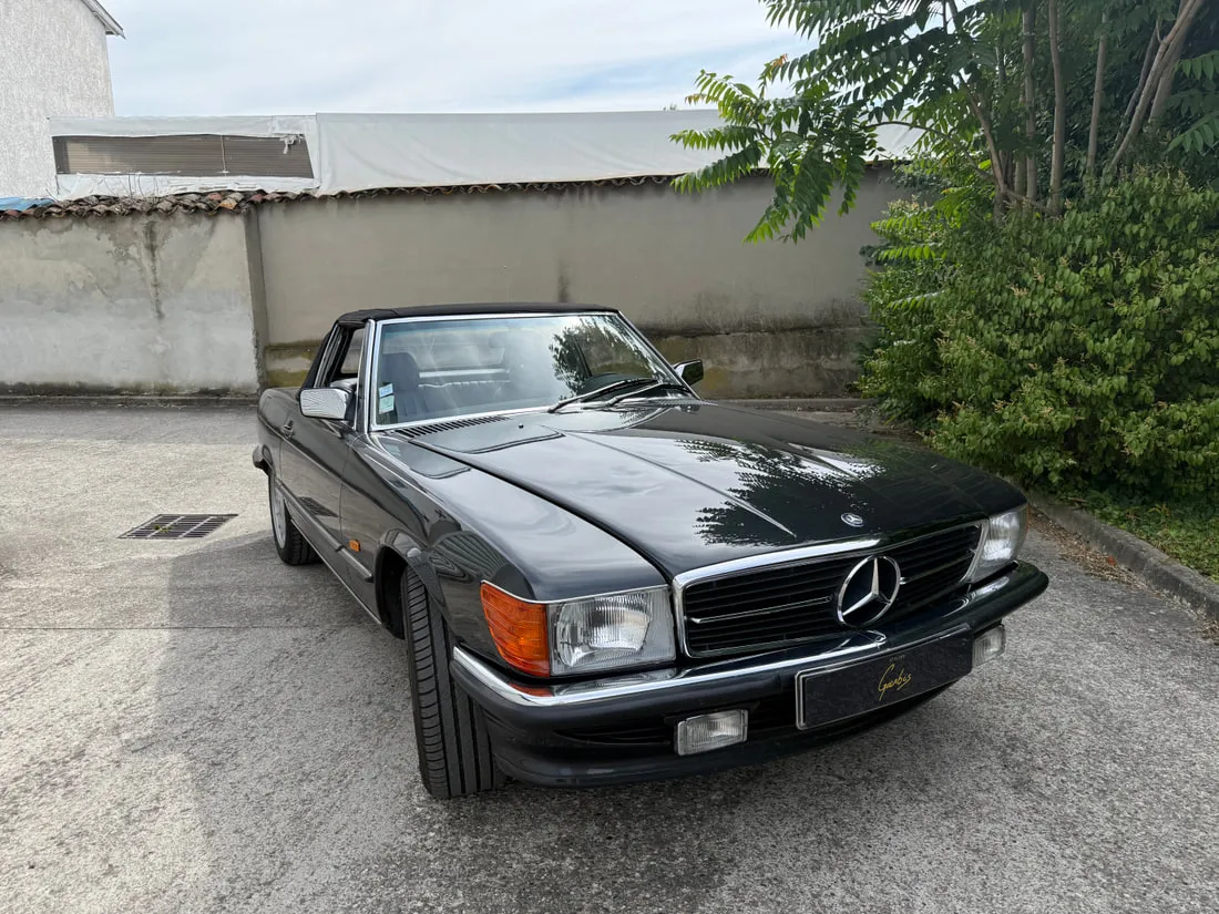 Mercedes R107 SL grise garée devant l’atelier, capote fermée, vue de trois quarts avant